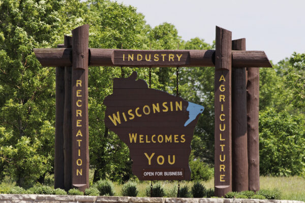 Large roadside sign shaped like Wisconsin reads, “Wisconsin Welcomes You – Open for Business,” with words “Industry,” “Recreation,” and “Agriculture” on surrounding wooden posts.