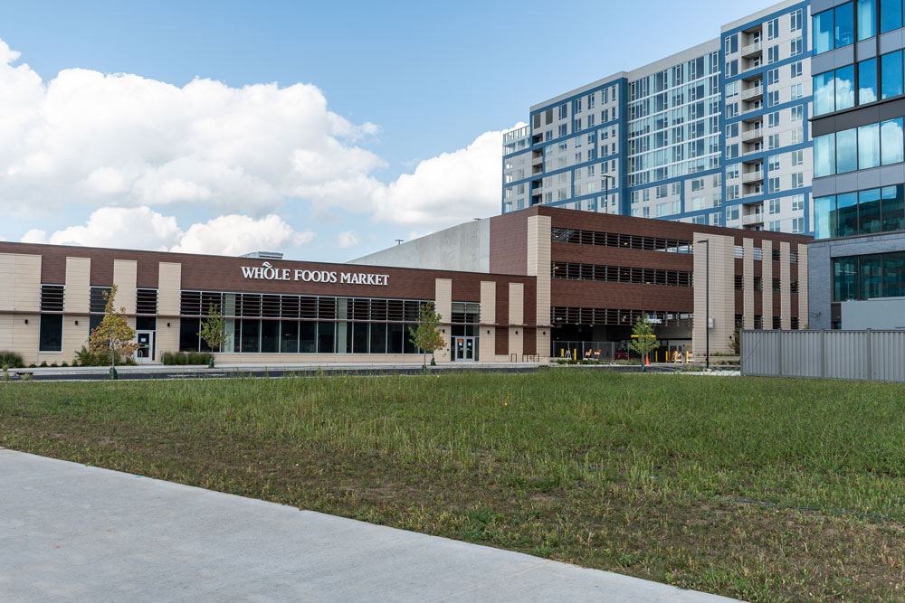 A Whole Foods Market storefront is attached to a modern multi-story building, with a grassy area and sidewalk in the foreground.