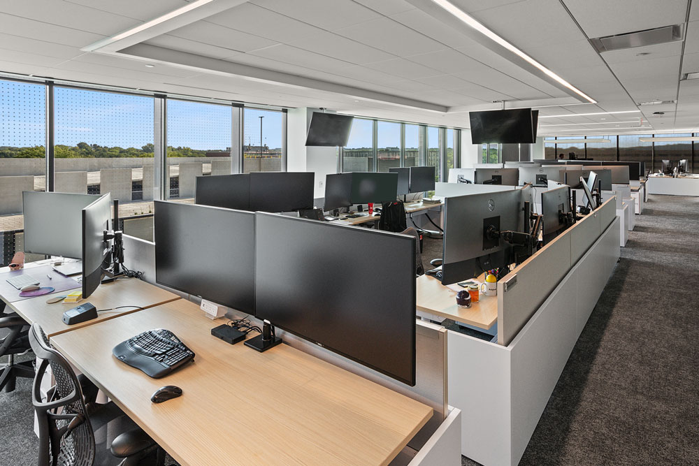 Modern open-plan office with rows of desks, multiple computer monitors, office supplies, and large windows allowing natural light.