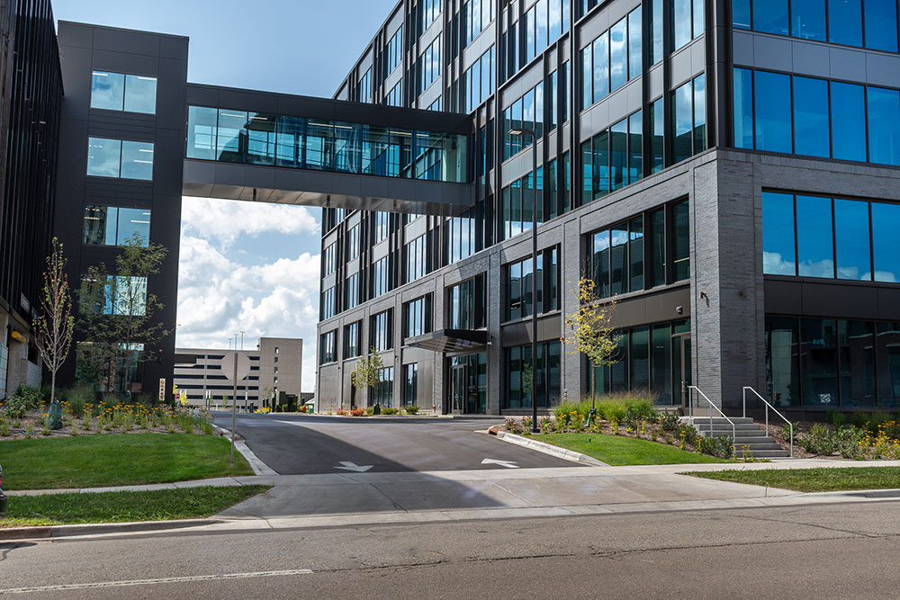 Modern office building with large glass windows, a skybridge connecting two sections, landscaping, and an empty driveway leading to the entrance.