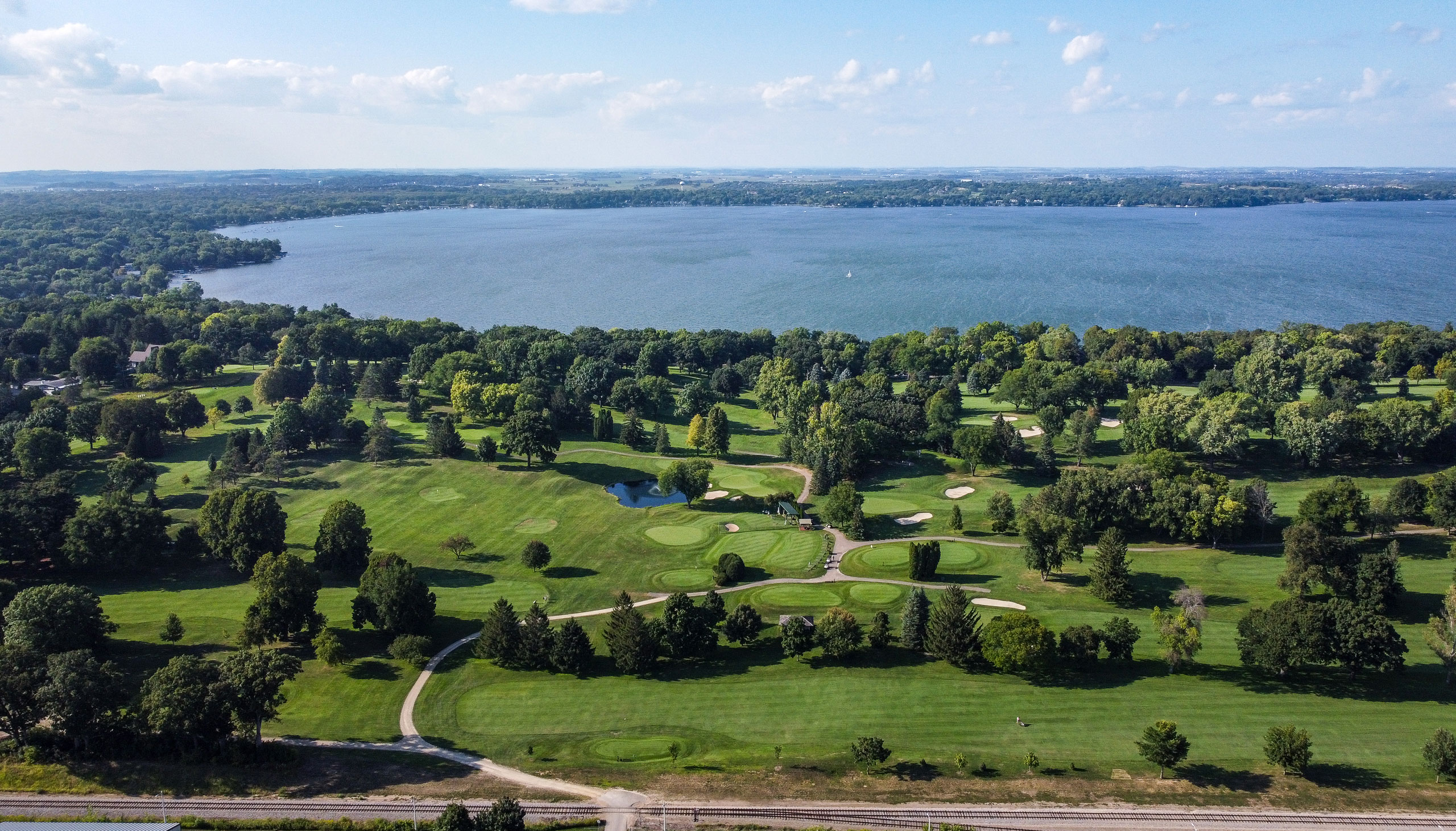 Aerial view of a golf course with green fairways and sand bunkers, bordered by trees, near a large lake under a partly cloudy sky.