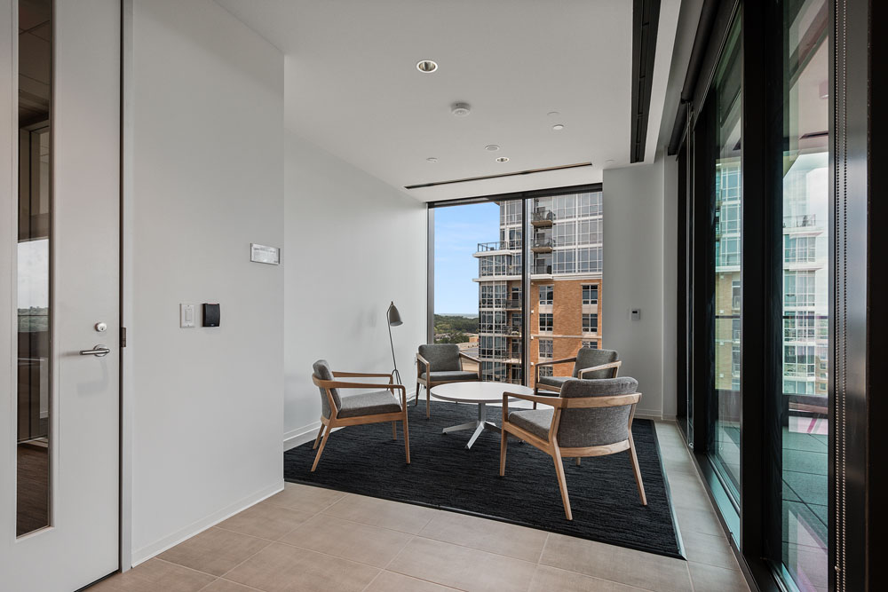 Small modern seating area with four chairs around a round table on a dark rug, next to large windows overlooking city buildings.