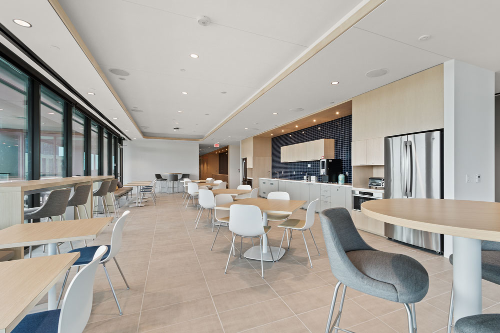 Modern office kitchen and break area with tables, chairs, bar seating, stainless steel appliances, large windows, and neutral color palette.