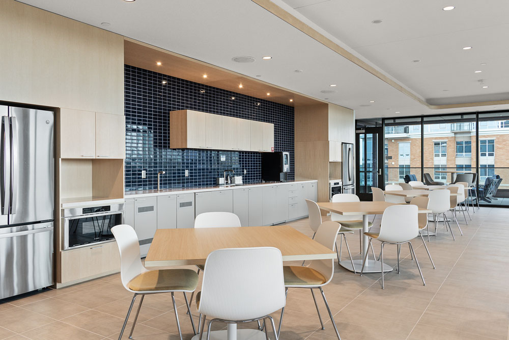 Modern office kitchen with stainless steel appliances, blue tile backsplash, light wood cabinets, and several tables with white chairs arranged in the dining area.