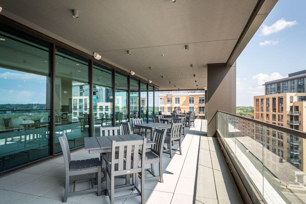 Outdoor terrace with several tables and chairs, glass railing, shaded ceiling, and city buildings visible in the background under a partly cloudy sky.