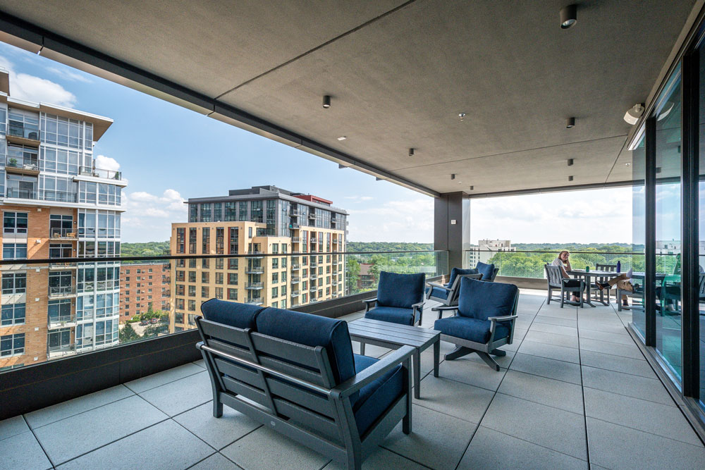 Modern outdoor terrace with blue cushioned seating and tables, overlooking city buildings and greenery, under a covered roof. Two people sit at a table in the background.