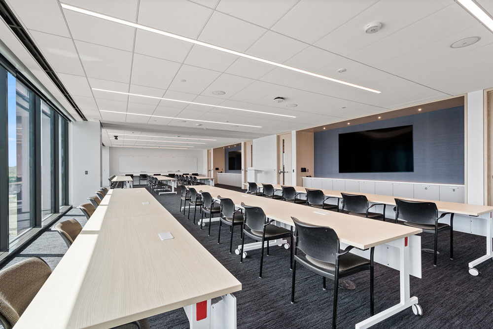 A modern conference room with several long tables, black chairs, large windows, a whiteboard, and a large wall-mounted screen at the front.