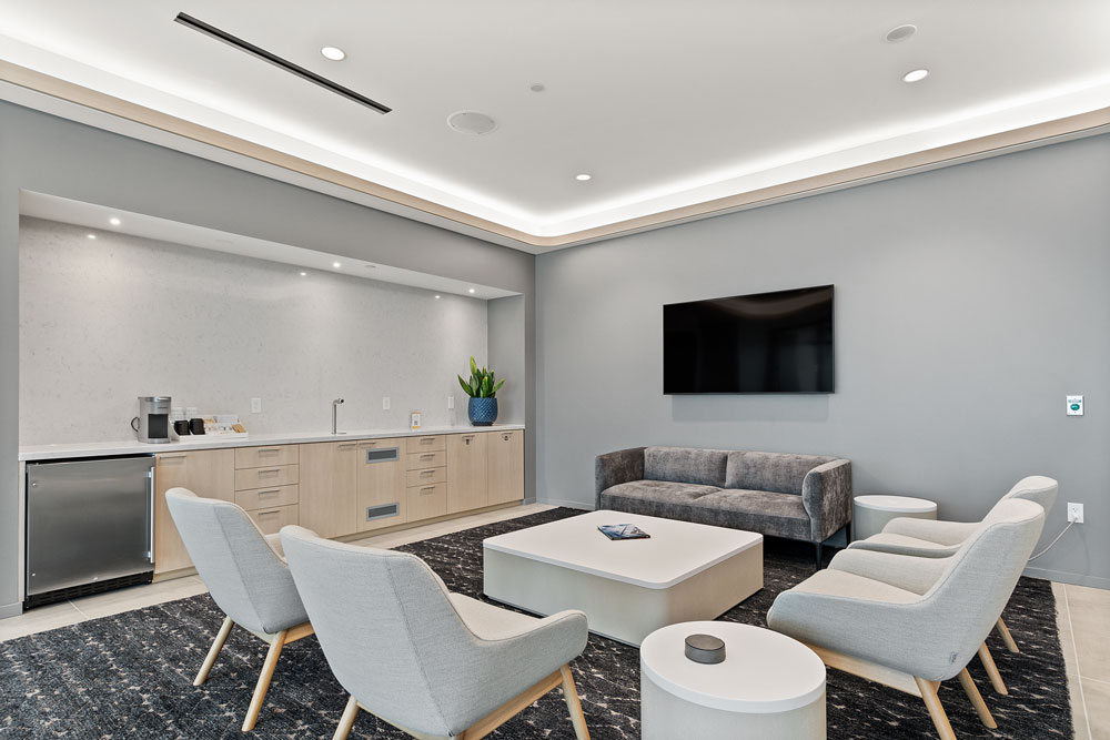 Modern lounge area with light gray chairs, a dark rug, a sofa, coffee table, wall-mounted TV, and a kitchenette with a sink and mini fridge.