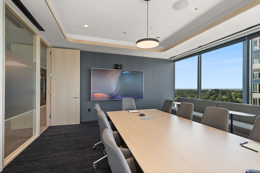 Modern conference room with a long wooden table, gray chairs, large wall-mounted screen, and floor-to-ceiling windows overlooking a cityscape and greenery.