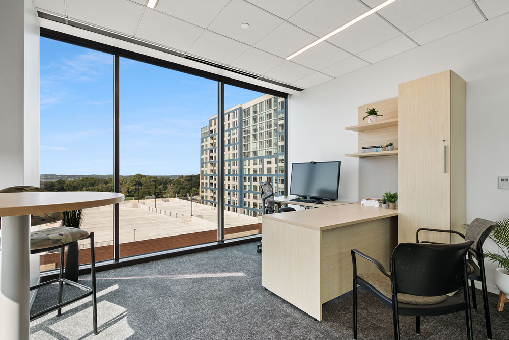 Modern office with light wood furniture, large window with city and nature view, desk with computer, chairs, shelves, and minimal decor.
