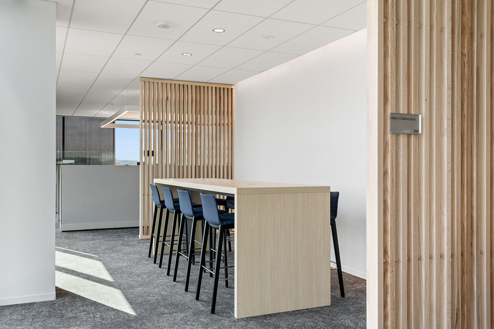 A modern office space with a tall wooden table, six blue barstools, slatted wood partitions, and a sign reading "Open Collaboration" on the wall.