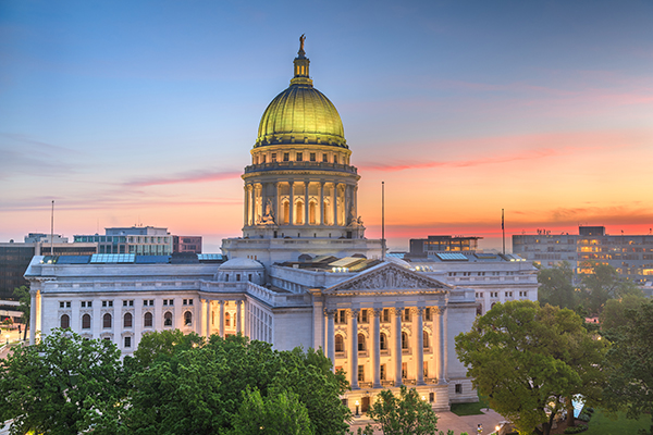 The Wisconsin State Capitol building with its domed roof is shown at sunset, with trees in the foreground and a colorful sky in the background.