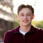 A young man with short light brown hair smiles at the camera outdoors, wearing a maroon zip-up sweater over a white shirt.