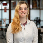 A young woman with glasses and long blonde hair, wearing a white zip-up jacket, stands indoors in a room with tables and blurred background.