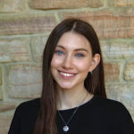 A young woman with long brown hair and blue eyes smiles at the camera, standing in front of a stone wall. She is wearing a black top and a necklace.