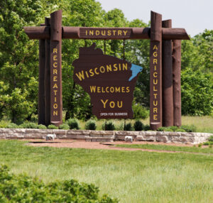 A wooden roadside sign reads "Wisconsin Welcomes You" with "Industry," "Agriculture," and "Recreation" on its frame, set among greenery and trees.